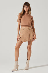 A woman is wearing a loose-fitting, short-sleeved, tan top that reaches just above her waist. She pairs it with a high-waisted, textured beige mini skirt featuring decorative buttons and zippered pockets on each side. She completes her outfit with shiny, cream-colored ankle boots with stiletto heels.